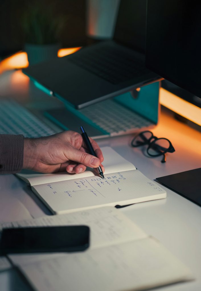 Hand writing a website checklist in a notebook beside a laptop